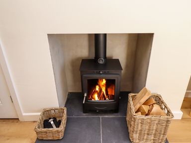 A wood burning stove with fire inside and two wicker baskets one with firewood the other with tools at 2 Twizel Station Cottage in Cornhill-on-Tweed near Norham
