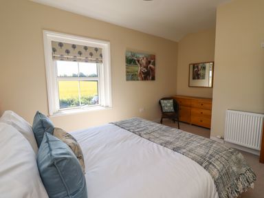 A bedroom with a bed pillows and a blanket a window with a patterned blind a wooden dresser a wicker chair with cushion a painting of a highland cow and a radiator at 2 Twizel Station Cottage Cornhill-on-Tweed near Norham