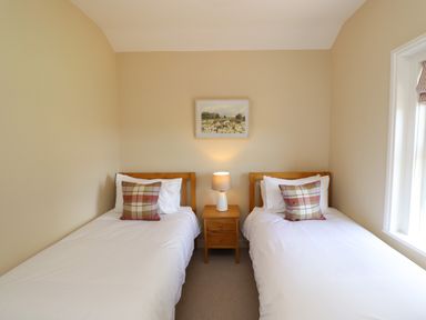 A bedroom with two single beds with plaid pillows and a wooden nightstand with a lamp between at 2 Twizel Station Cottage in Cornhill-on-Tweed near Norham