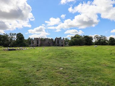 A grassy field with sheep grazing and an old stone ruin in the background surrounded by trees