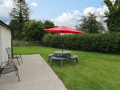 A patio with two metal chairs and a round picnic table with a red umbrella on a lawn at Marys Cottage Theur near Riverstown County Sligo