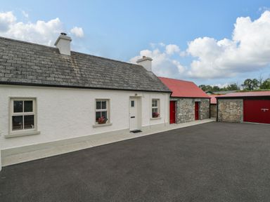 An exterior view of a white cottage with gray roof and red doors next to stone buildings with red roofs at Marys Cottage Theur near Riverstown County Sligo
