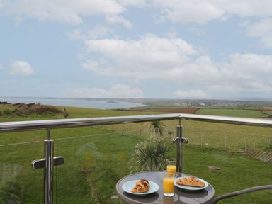 A balcony with a table holding two plates with croissants and two glasses of orange juice overlooking green fields and a coastline at 5 Ocean Reach in Bolberry