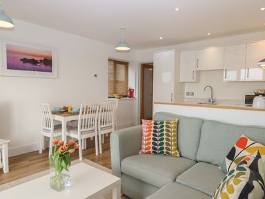 An open living area with a light blue sofa with patterned cushions a dining table with four white chairs kitchen cabinets and a vase of tulips on a white coffee table at 5 Ocean Reach in Bolberry