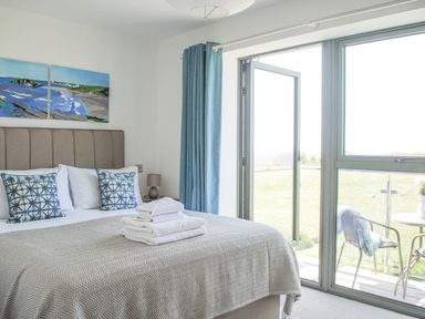 A bedroom with a bed with pillows and folded towels next to a window with blue curtains and a door leading to an outdoor seating area at 5 Ocean Reach in Bolberry