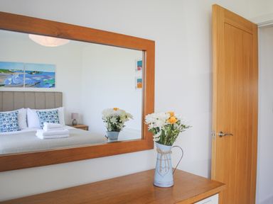 A wooden dresser with a vase of flowers and a mirror reflecting a bed with pillows and artwork at 5 Ocean Reach in Bolberry