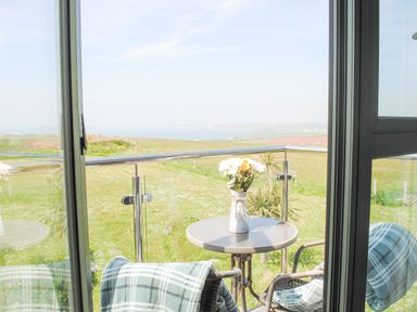 A balcony with a round table holding a vase of flowers and two chairs with plaid blankets overlooking green fields and distant water at 5 Ocean Reach in Bolberry