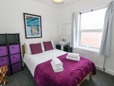 A bedroom with a bed covered in purple bedding and white towels on top a black storage unit a sink below a mirror and a window with curtains at Beachaven in Weymouth