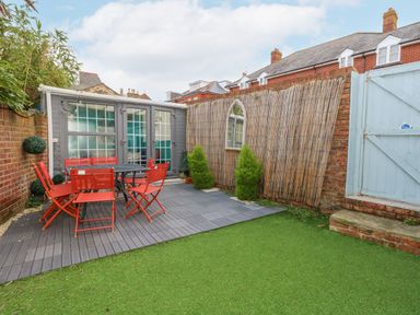 A small backyard with red chairs around a black table on a gray deck and artificial grass at Beachaven in Weymouth