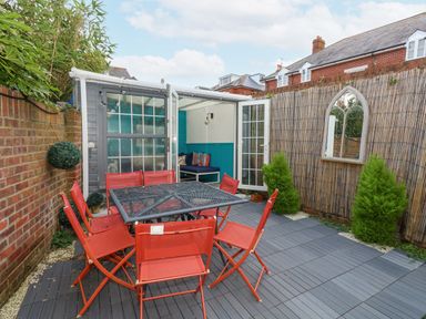 A backyard patio with red chairs around a black table a small garden room with glass doors and green plants at Beachaven in Weymouth