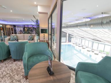 A lounge area with teal armchairs and a round wooden table next to a glass window overlooking an indoor swimming pool with lounge chairs at Classic Lodge 25 in Dyserth
