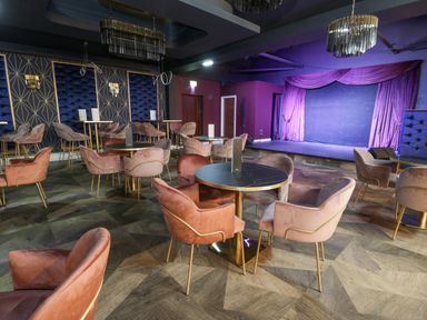A dining area with round tables and pink velvet chairs facing a stage with purple curtains at Classic Lodge 25 in Dyserth