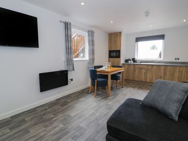 A kitchen and dining area with wooden cabinets a wooden table and blue chairs a gray sofa and a wall-mounted television at Classic Lodge 25 in Dyserth