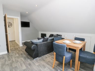 A dining area with a wooden table and four blue chairs next to a living space with two black sofas and a wall-mounted television at Classic Lodge 15 in Dyserth