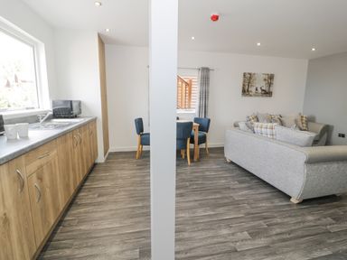 A kitchen with wooden cabinets and a dining area with blue chairs next to a living room with grey sofas at Luxury Lodge 46 in Dyserth