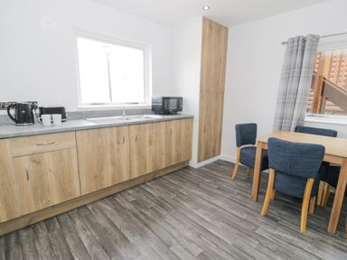 A kitchen with wooden cabinets a gray countertop a sink a microwave kettle and cups a wooden dining table with blue chairs and gray curtains at Luxury Lodge 46 in Dyserth
