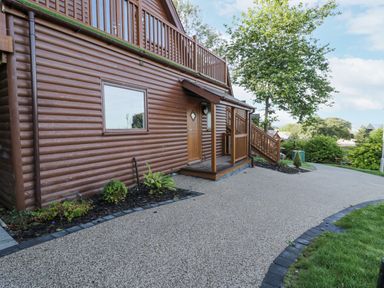 A brown wooden lodge with a small porch and stairs surrounded by greenery and a paved driveway at Luxury Lodge 46 in Dyserth