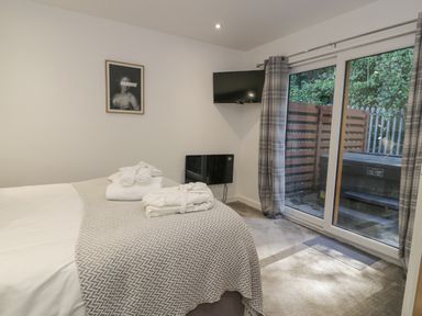 A bedroom with a bed covered with a blanket towels and bathrobe a wall mounted tv and sliding glass doors with curtains overlooking an outdoor area at Luxury Lodge 46 in Dyserth