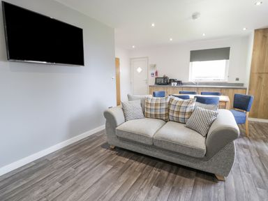 A living room with a grey sofa with cushions a wall-mounted television wooden dining table with blue chairs and a kitchenette at Luxury Lodge 62 in Dyserth