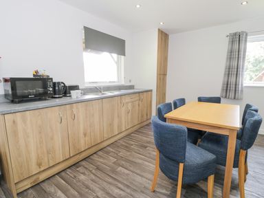 A kitchen with wooden cabinets a countertop with a microwave kettle cups a dining table with six blue upholstered chairs and two windows with blinds and curtains at Luxury Lodge 62 in Dyserth