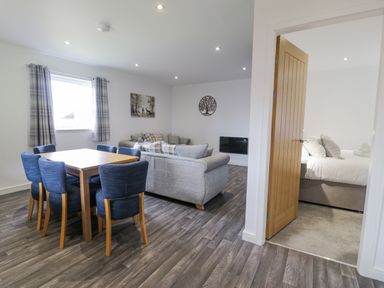 A dining area with a wooden table and six blue chairs next to a living room with grey sofas and a wall-mounted TV at Luxury Lodge 62 in Dyserth