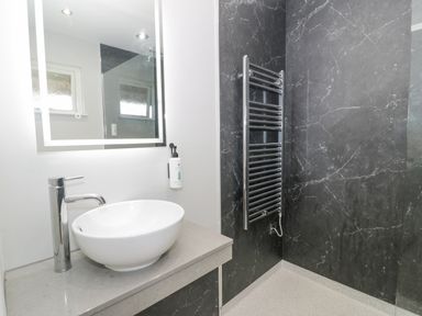 A bathroom with a white round sink on a countertop a chrome faucet a backlit mirror a soap dispenser and a chrome towel radiator on a dark marble wall at Luxury Lodge 62 in Dyserth