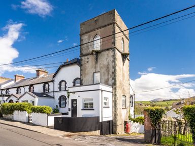A street view of a tall stone tower attached to a white house with black trim and nearby greenery at The Old Coastguard Tower Buncrana Buncrana County Donegal