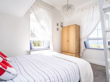 A bedroom with a bed striped bedding a wooden wardrobe and two windows with seating at The Old Coastguard Tower Buncrana in Buncrana County Donegal