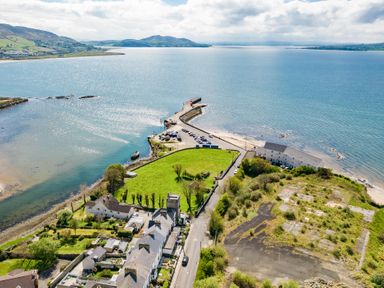 A coastal area with buildings, roads, green fields, a pier with parked cars, and surrounding water at The Old Coastguard Tower Buncrana, County Donegal