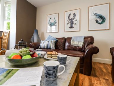 A living room with a brown leather sofa three framed sea creature pictures and a coffee table with mugs fruit and pastries at The Lobster Pot in Weymouth