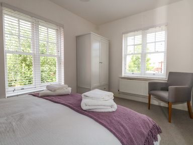 A bedroom with a bed, folded towels on the bed, a white wardrobe, a grey armchair, and two windows with blinds at The Lobster Pot in Weymouth