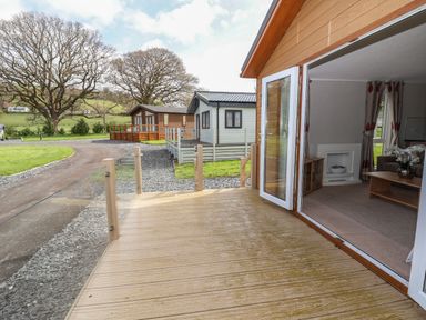 A wooden deck outside a house with an open door showing a living room with curtains and furniture a pathway a grassy area and two other similar houses in the background at Lodge 4 in St Asaph