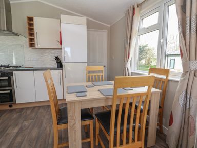 A dining area with a wooden table and four chairs next to a kitchen with white cabinets and a refrigerator at Lodge 4 in St Asaph