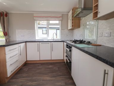 A kitchen with white cabinets, a black countertop, a gas stove, an oven, a window with a blind, and a wooden floor at Lodge 4 in St Asaph
