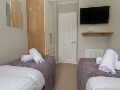 A bedroom with two single beds with towels on top a wardrobe a wall mounted tv and a door at Lodge 4 in St Asaph