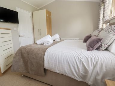 A bedroom with a bed with pillows and towels a wardrobe a dresser and a wall mounted television at Lodge 4 in St Asaph