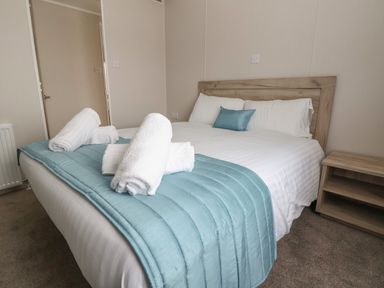 A bedroom with a double bed with white bedding and blue throw and pillows with rolled white towels on the bed and a wooden nightstand at Lodge 9 in St Asaph