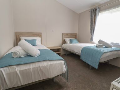 A bedroom with two single beds blue bed runners and pillows a small wooden nightstand between the beds and a window with curtains at Lodge 9 in St Asaph