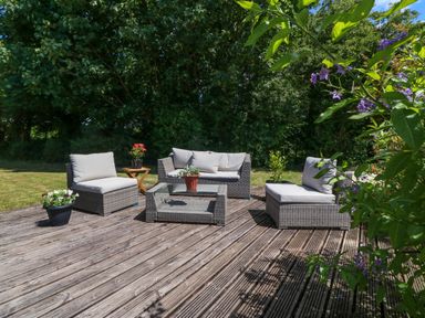 An outdoor wooden deck with woven patio furniture and potted plants surrounded by trees and grass at Ty Gareg in Llanddaniel near Gaerwen