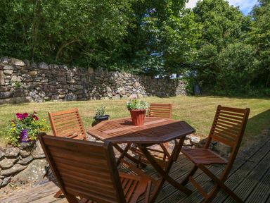 A garden area with a wooden table and chairs on a deck surrounded by grass and stone wall at Ty Gareg in Llanddaniel near Gaerwen