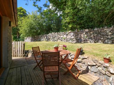 An outdoor wooden deck with a wooden table and four chairs with plants on the table and stone walls in a garden at Ty Gareg in Llanddaniel near Gaerwen