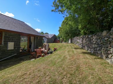 A grassy yard with a wooden table and chairs next to a stone house and a stone wall with trees at Ty Gareg in Llanddaniel near Gaerwen