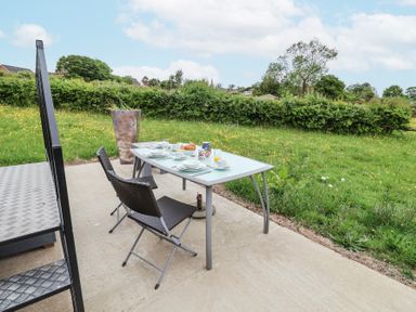 An outdoor dining table with two chairs on a patio next to a grassy field with trees at The Hoot in Great Finborough near Stowmarket
