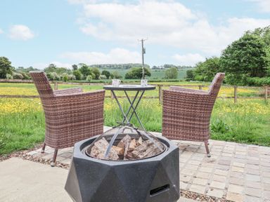 An outdoor patio with two wicker chairs a small round table with mugs and a fire pit with wood at The Hoot in Great Finborough near Stowmarket