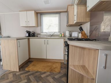 A kitchen with white and wood cabinets a window with blinds a stove a kettle a coffee machine and a brown mat on the floor at Woodlands Lodge in Bontuchel near Ruthin
