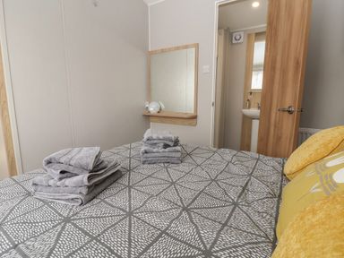 A bedroom with a patterned bedspread folded towels on the bed a wall-mounted mirror and an open door leading to a bathroom at Woodlands Lodge in Bontuchel near Ruthin