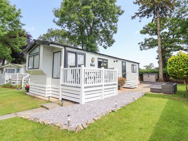 A white mobile home with a small deck and outdoor seating on a grass lawn surrounded by trees at Woodlands Lodge in Bontuchel near Ruthin