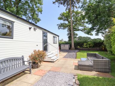 An outdoor patio area with a bench a potted plant a white mobile home wicker furniture grass trees and a storage shed at Woodlands Lodge in Bontuchel near Ruthin