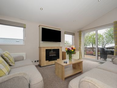 A living room with two sofas a coffee table with flowers a mounted television and sliding glass doors at Lodge 67 at Riviera Bay Coastal Retreat Brixham