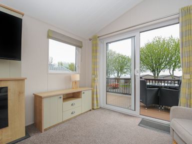 A living area with a cabinet and lamp beside a window and sliding glass door leading to a balcony with chairs at Lodge 67 at Riviera Bay Coastal Retreat in Brixham
